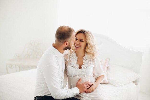 Retrato de una hermosa pareja de mamá y papá abrazando el estómago y mirándose tocándose cara a cara Mujer rubia embarazada de blanco sonriendo a su apuesto esposo posando en un elegante interior