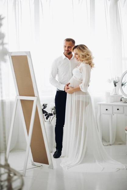 Retrato de una hermosa pareja de mamá y papá abrazando el estómago y mirándose tocándose cara a cara Mujer rubia embarazada de blanco sonriendo a su apuesto esposo posando en un elegante interior