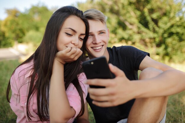 Retrato de una hermosa pareja sonriente sentada en el césped en el parque y felizmente usando el teléfono móvil juntos