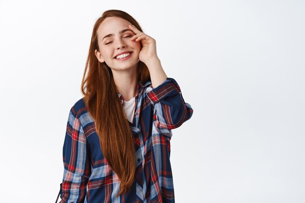 Retrato de una hermosa pelirroja con cabello largo natural, ojos cerrados y sonriente, tocando la cara sin maquillaje, piel pura y suave, de pie sobre fondo blanco