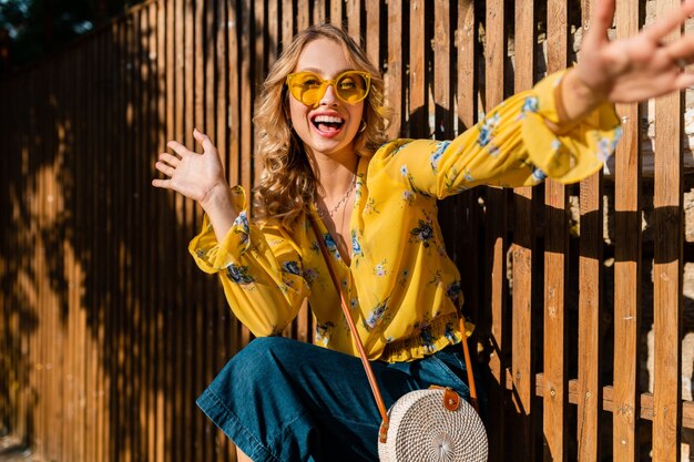 Retrato de hermosa rubia emocional riendo elegante mujer sonriente en blusa amarilla con gafas de sol, bolso de paja estilo bali