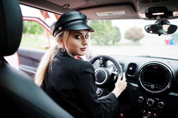 Retrato de hermosa rubia sexy modelo de mujer de moda en gorra y en todo negro con maquillaje brillante sentarse y conducir coche rojo de la ciudad