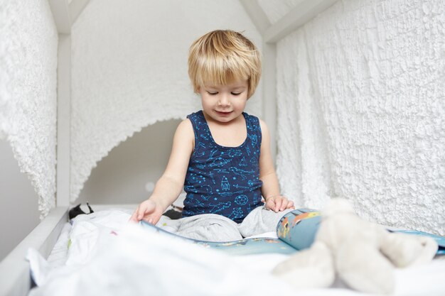 Retrato de un hermoso bebé caucásico con cabello rubio vestido con pijama sentado en una cama con dosel blanco, absorto en la lectura de libros para niños, mirando a través de imágenes con expresión interesada