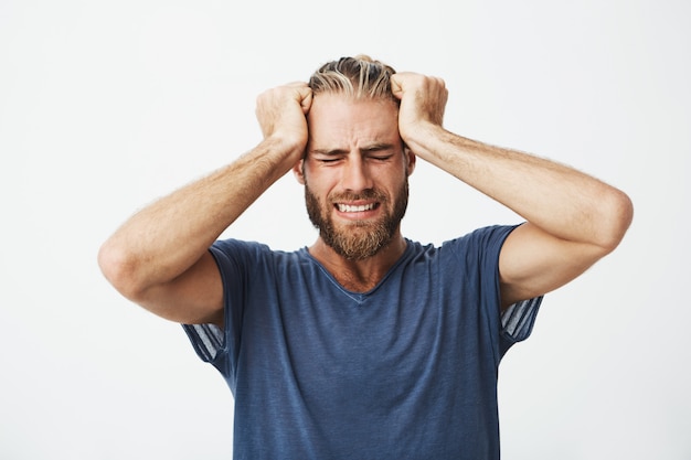 Foto gratuita retrato de hermoso chico nórdico con corte de pelo moderno y barba sosteniendo la cabeza con las manos sufriendo de dolor de cabeza