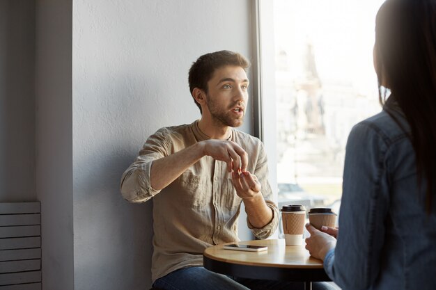 Retrato del hermoso diseñador independiente sin afeitar, sentado en un café en una reunión con un cliente que intenta explicar la concepción de su trabajo y expresivo gesticulando con las manos.