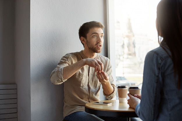 Retrato del hermoso diseñador independiente sin afeitar, sentado en un café en una reunión con un cliente que intenta explicar la concepción de su trabajo y expresivo gesticulando con las manos.