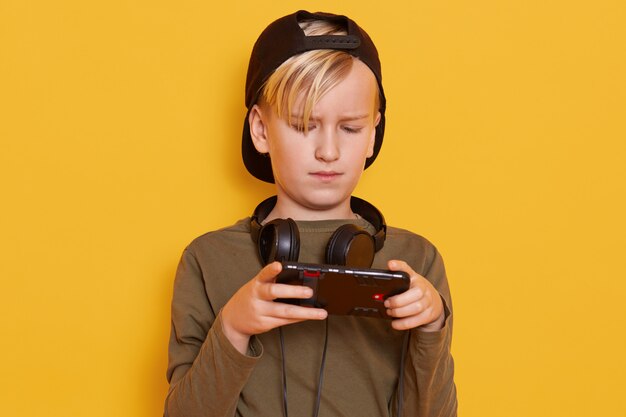 Retrato de un hermoso y pequeño chico rubio, con una mirada concentrada y seria mientras usa el teléfono móvil, niño jugando videojuegos en línea