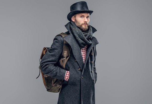 Retrato de un hipster barbudo vestido con una chaqueta gris y un sombrero cilíndrico sostiene una mochila aislada en un fondo gris.