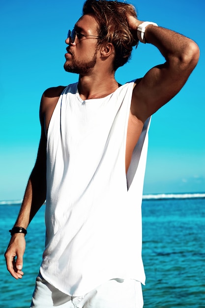 Retrato de hipster guapo modelo de hombre de moda tomando el sol con ropa casual en camiseta blanca y gafas de sol posando en el océano azul y el cielo