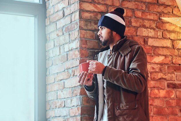 Retrato de un hipster indio barbudo vestido con una chaqueta cálida y un sombrero, bebe café de una taza roja cerca de la pared de un ladrillo rojo.