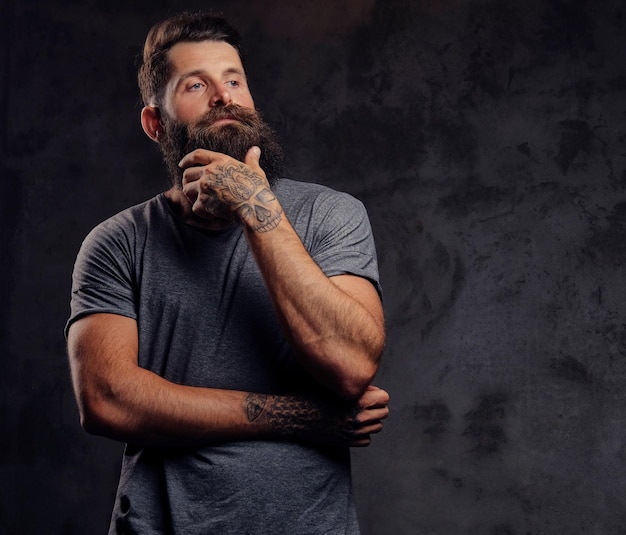 Retrato de un hipster tatuado con barba completa y corte de pelo elegante, vestido con una camiseta gris, se para con una mirada pensante en un estudio sobre un fondo oscuro.