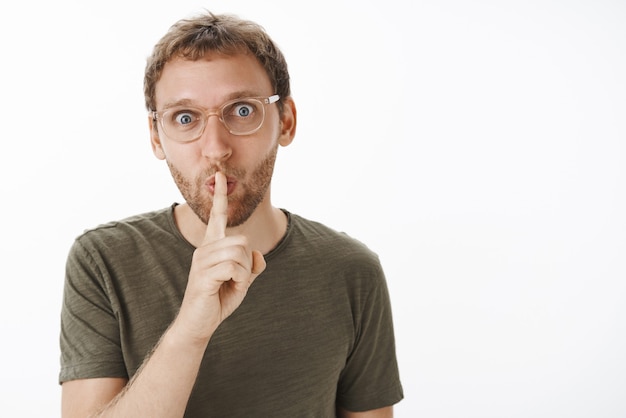 Retrato de hombre adulto atractivo, divertido y alegre entusiasta con labios plegables de cerdas en sonido shhh sosteniendo el dedo índice sobre la boca mientras calla para guardar el secreto