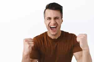 Foto gratuita retrato de un hombre adulto guapo triunfante cantando celebrando el éxito o el logro apretando los puños y gritando sí con una sonrisa radiante y feliz ganando el premio convertido en campeón viendo un juego deportivo