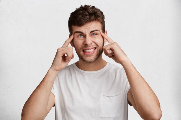 Retrato de hombre sin afeitar estresante infeliz viste camiseta blanca, mantiene los dedos en las sienes, aprieta los dientes, aislado sobre fondo de estudio. Hombre barbudo joven disgustado expresa emociones negativas