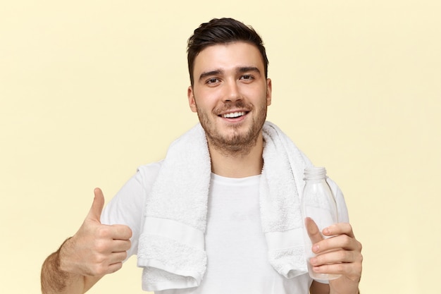 Retrato de hombre sin afeitar joven alegre guapo con amplia sonrisa de confianza haciendo pulgares arriba gesto refrescándose después de entrenar en el gimnasio