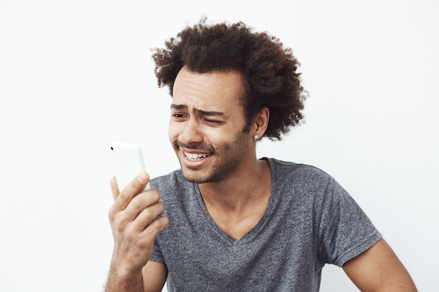 Retrato de hombre africano confundido pero alegre y guapo sonriendo mirando el teléfono celular sorprendido con una foto en las redes sociales o una videollamada.