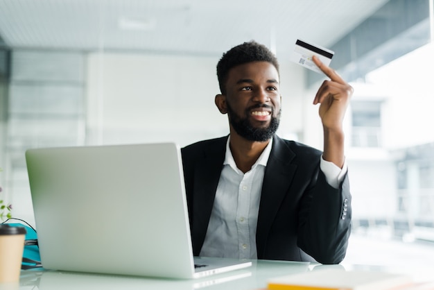 Retrato del hombre africano joven confidente que sostiene la tarjeta de crédito con la computadora portátil que paga vía Internet