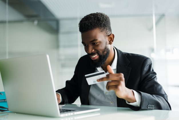 Retrato del hombre africano joven confidente que sostiene la tarjeta de crédito con la computadora portátil que paga vía Internet