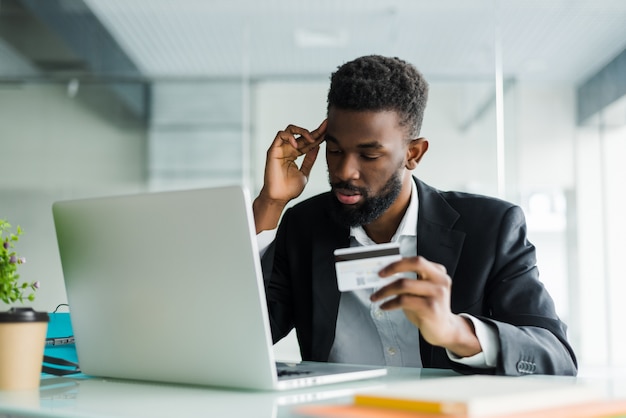 Retrato del hombre africano joven confidente que sostiene la tarjeta de crédito con la computadora portátil que paga vía Internet