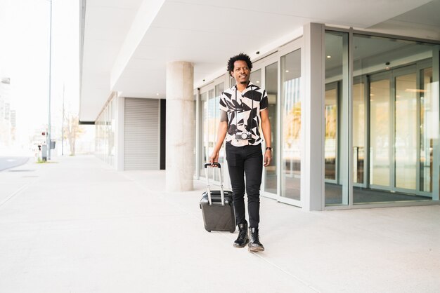 Retrato de hombre afro turista llevando maleta mientras camina al aire libre en la calle