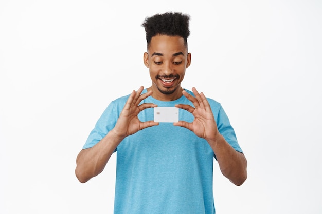 Retrato de un hombre afroamericano alegre que muestra una tarjeta de crédito, sonríe y se ve complacido con el descuento, parado en una camiseta azul contra un fondo blanco