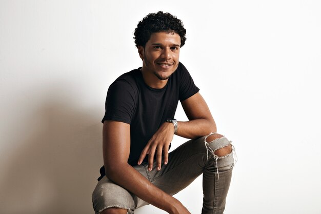 Retrato de un hombre afroamericano en una camiseta de algodón negro y jeans rotos sentado en la pared de un blanco