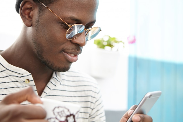 Retrato de hombre afroamericano con estilo moderno en ropa de moda disfrutando de conexión inalámbrica a internet gratuita en la cafetería, tomando café y leyendo mensajes en línea mientras pasa vacaciones en el extranjero