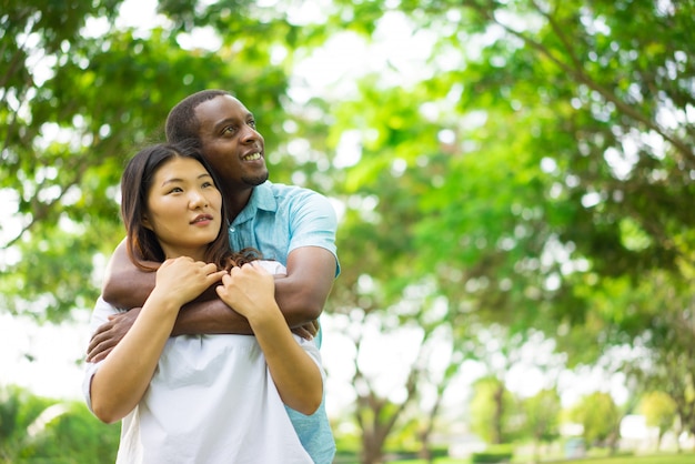 Retrato del hombre afroamericano feliz que abraza a la novia asiática y que mira lejos al aire libre