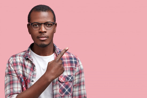 Retrato de hombre afroamericano guapo serio con expresión seria, usa gafas y camisa a cuadros, indica con el dedo índice en el espacio de copia en blanco para su