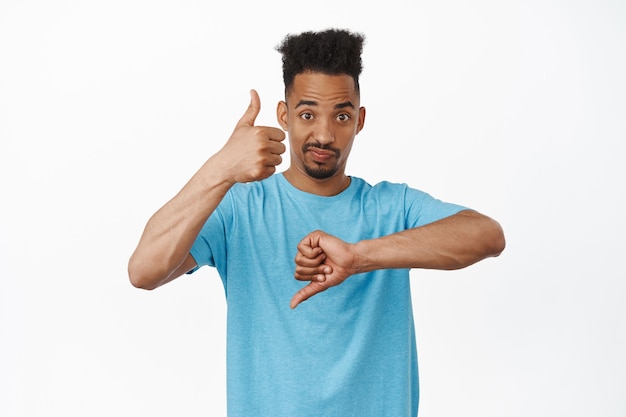 Foto gratuita retrato de hombre afroamericano mostrando los pulgares hacia arriba y hacia abajo, juzgando algo, tasa media cosa media, frunciendo el ceño perplejo para responder, de pie sobre blanco