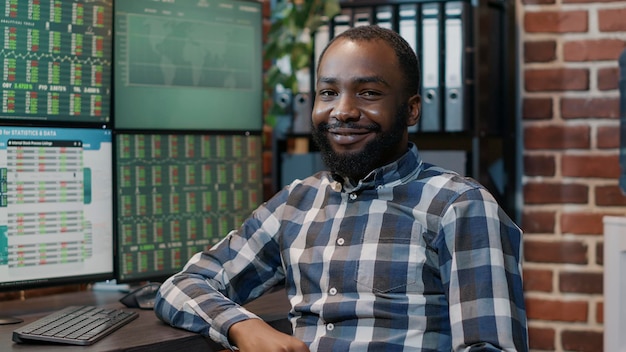 Retrato de un hombre afroamericano que usa monitores múltiples para trabajar en estrategia financiera con gráficos bursátiles y estadísticas de ventas de divisas. Participación de inversión con intercambio de capital y comercio de dinero.