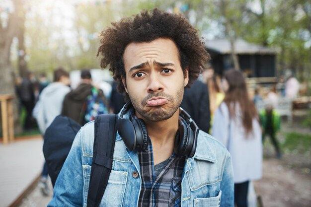 Retrato de hombre afroamericano sombrío con expresión linda, peinado afro y auriculares sobre el cuello, frunciendo el ceño mientras está molesto, de pie en el parque.