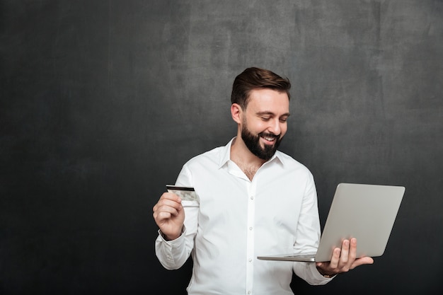 Retrato de hombre alegre haciendo pagos en línea en internet con notebook y tarjeta de crédito, aislado sobre gris oscuro