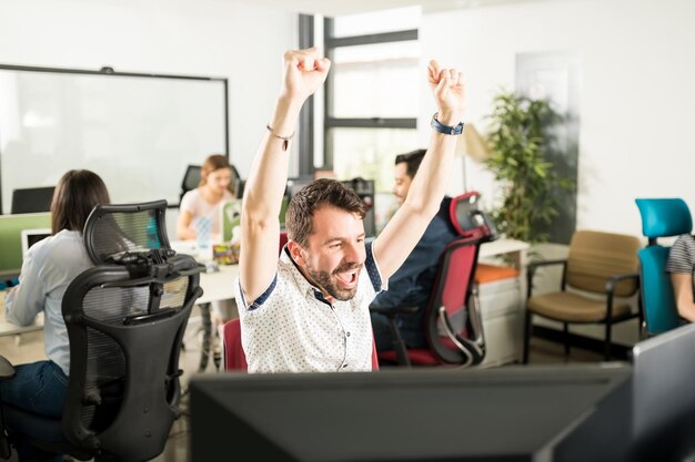 Retrato de un hombre alegre y positivo con atuendo causal levantando la mano y celebrando el logro sentado en una oficina moderna con colegas en el fondo