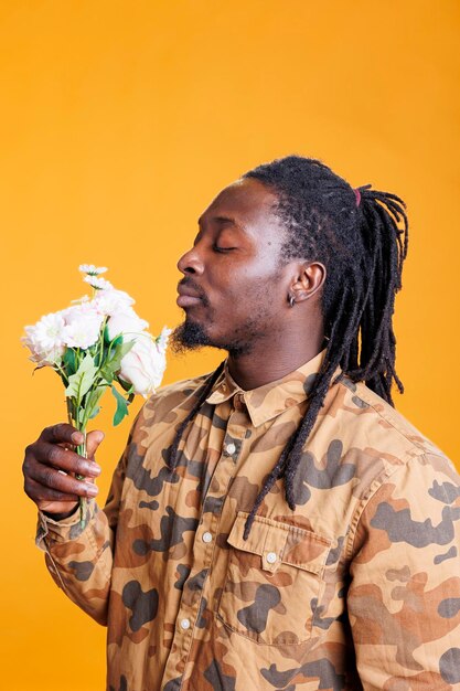 Retrato de un hombre alegre que disfruta del olor a rosas blancas, posando con flores para el día de San Valentín en un estudio sobre fondo amarillo. Adulto joven romántico afroamericano celebrando vacaciones de amor