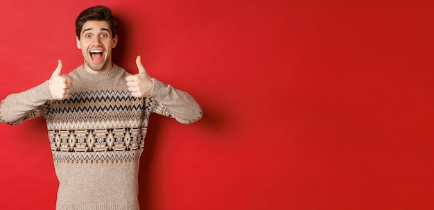 Retrato de un hombre alegre con suéter navideño como algo bueno, mostrando el pulgar hacia arriba y sonriendo asombrado, alabando la fiesta de año nuevo, de pie sobre un fondo rojo.