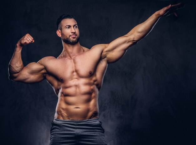 Retrato de un hombre alto y sin camisa con un cuerpo musculoso con un elegante corte de pelo y barba, en pantalones cortos deportivos, posando en un estudio. Aislado en un fondo oscuro.