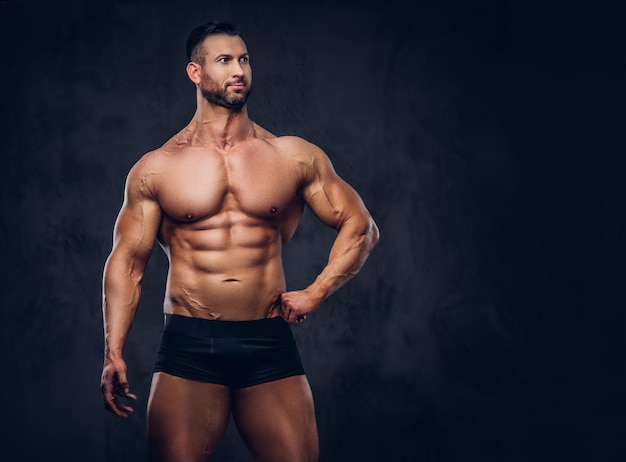 Retrato de un hombre alto y sin camisa con un cuerpo musculoso con un elegante corte de pelo y barba, en ropa interior, posando en un estudio. Aislado en un fondo oscuro.