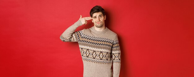 Retrato de un hombre apuesto angustiado y molesto, vestido con un suéter de Navidad, haciendo un signo de pistola con el dedo cerca de la cabeza y disparándose a sí mismo, de pie sobre un fondo rojo