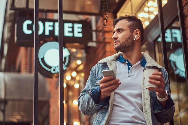 Retrato de un hombre apuesto y elegante con una chaqueta de denim con auriculares inalámbricos sosteniendo café para llevar fuera de la cafetería.