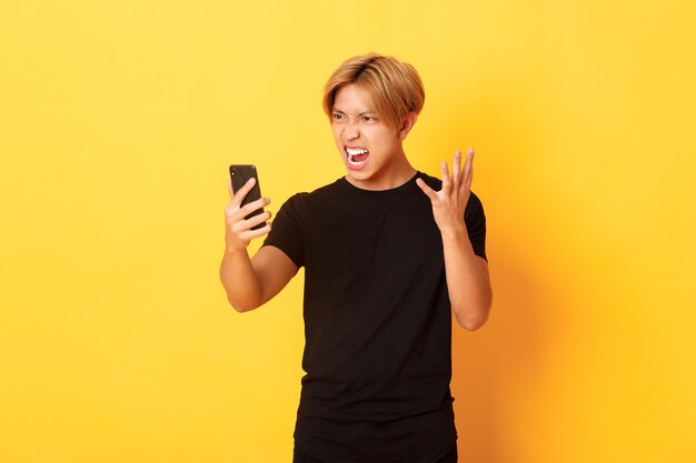 Retrato de hombre asiático enojado y cabreado mirando enojado con la pantalla del teléfono inteligente, teniendo una discusión durante la videollamada, pared amarilla de pie