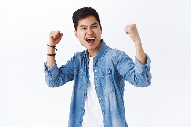 Retrato de un hombre asiático entusiasta empoderado que levanta las manos en triunfo celebrando la victoria sonriendo diciendo que sí ganar el juego o la competencia lograr el objetivo de fondo blanco