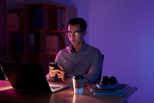 Retrato de hombre asiático enviando mensajes de texto en el cuarto oscuro sentado en la computadora portátil