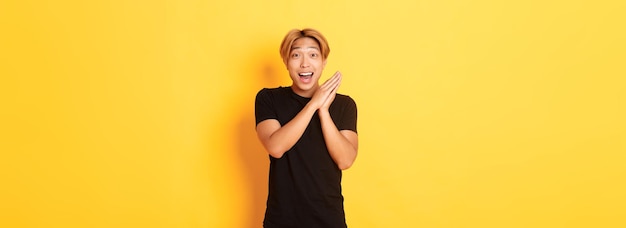 Retrato de un hombre asiático feliz y sorprendido aplaudiendo y sonriendo encantado de pie fondo amarillo