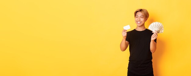 Retrato de un hombre asiático guapo sonriente satisfecho que se ve complacido con la tarjeta de crédito mientras también sostiene m