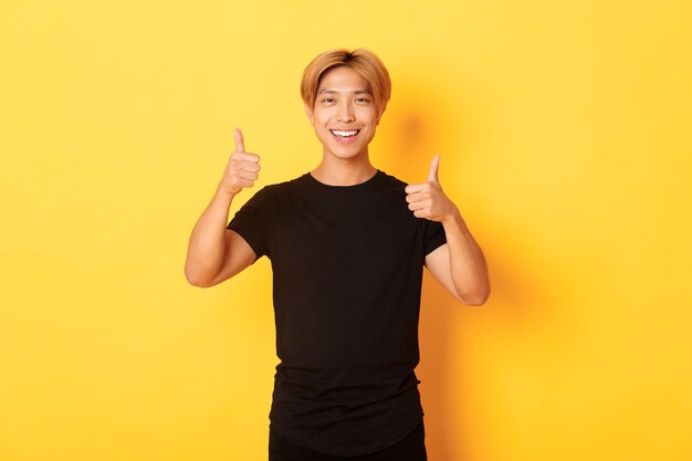 Retrato de hombre asiático satisfecho sonriendo y mostrando el pulgar hacia arriba sobre la pared amarilla