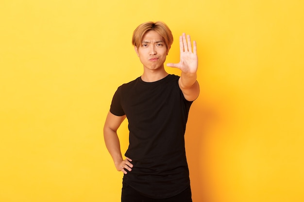 Retrato de hombre asiático serio decepcionado, sonriendo disgustado y extendiendo la mano, mostrando gesto de parada, pared amarilla