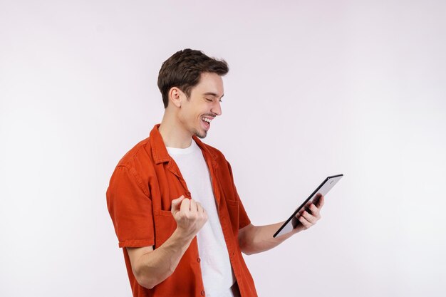 Retrato de un hombre atractivo y alegre haciendo un gesto de ganador apretando el puño y usando una aplicación de dispositivo buscando en la web aislada sobre un fondo de color blanco