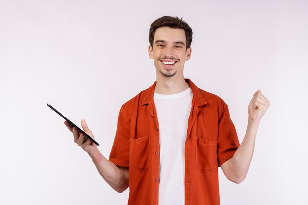 Retrato de un hombre atractivo y alegre haciendo un gesto de ganador apretando el puño y usando una aplicación de dispositivo buscando en la web aislada sobre un fondo de color blanco