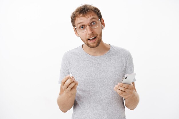 Retrato de hombre atractivo despreocupado cuestionado con barba inclinando la cabeza quitándose el auricular inalámbrico y sosteniendo el teléfono inteligente escuchando música